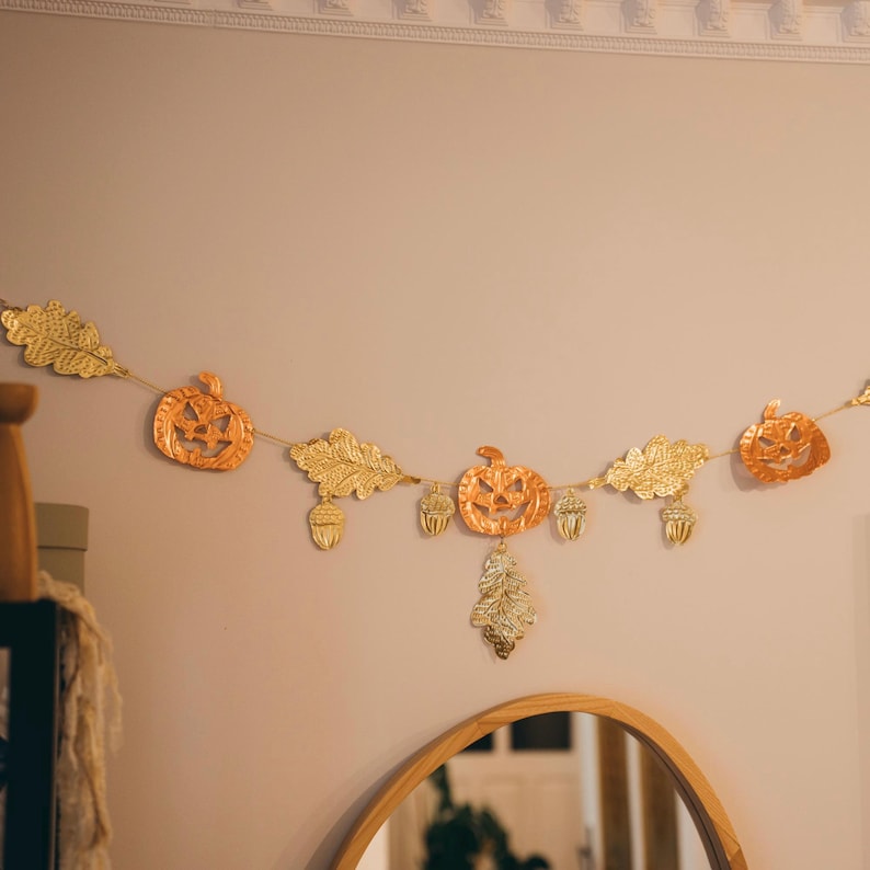 May include: A festive Halloween garland featuring orange pumpkin cutouts, gold-colored leaves, and acorns. The garland is strung on a thin gold cord and is displayed against a neutral wall. The garland is a decorative item for the fall season.
