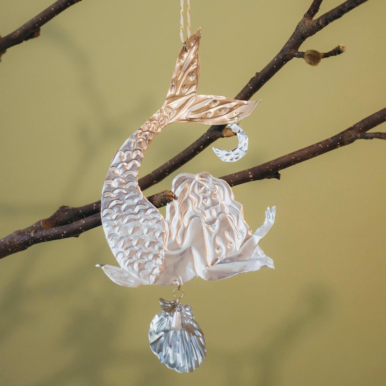 May include: A silver mermaid ornament with a gold tail and a seashell charm. The mermaid is hanging from a tree branch. The ornament has a detailed design and is made of a shiny material.