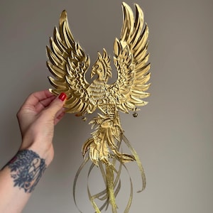 May include: A golden phoenix sculpture with outstretched wings and detailed feathering. The bird has a detailed head and tail, with flowing ribbons hanging from the bottom. The sculpture is held by a hand with red nail polish.