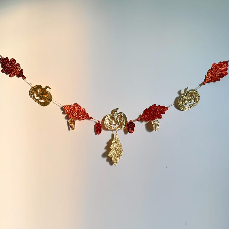 May include: A festive garland featuring gold pumpkins, red leaves, and gold acorns. The garland is strung on a thin white string and is perfect for fall or Halloween decorations. The decorative elements are made of a shiny material.