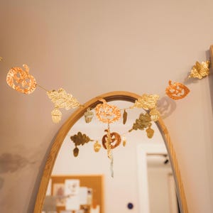 May include: A gold-colored autumn-themed garland with cut-out pumpkin, leaf, and acorn shapes. The garland is strung across a room, reflecting in a mirror. The garland is a decorative item for fall or Halloween.