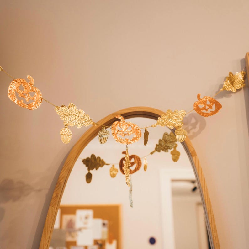 May include: A gold-colored autumn-themed garland with cut-out pumpkin, leaf, and acorn shapes. The garland is strung across a room, reflecting in a mirror. The garland is a decorative item for fall or Halloween.