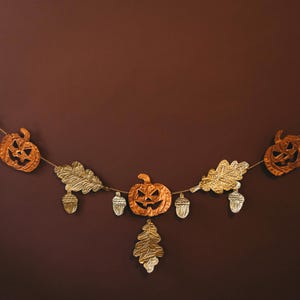May include: A decorative Halloween garland featuring orange jack-o'-lanterns, gold oak leaves, and acorns. The garland is strung on a thin, natural-colored cord, creating a festive autumn decoration. The background is a solid, dark brown.