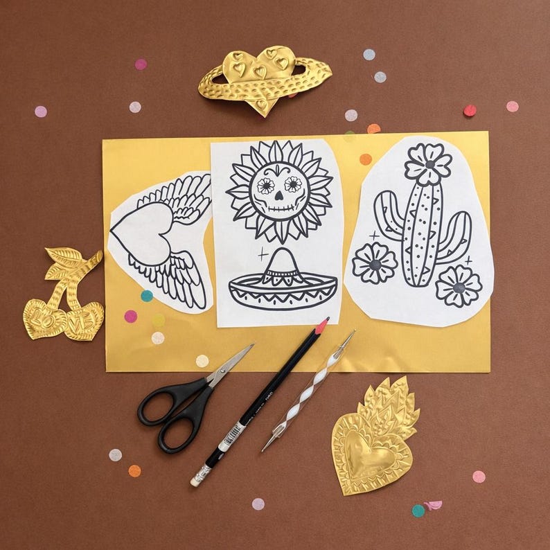 May include: A collection of craft supplies and decorative elements arranged on a brown surface. Includes gold-colored metal charms in the shape of a cherry, heart, and a crown. There are also black and white line drawings of a heart with wings, a sunflower skull, and a cactus.