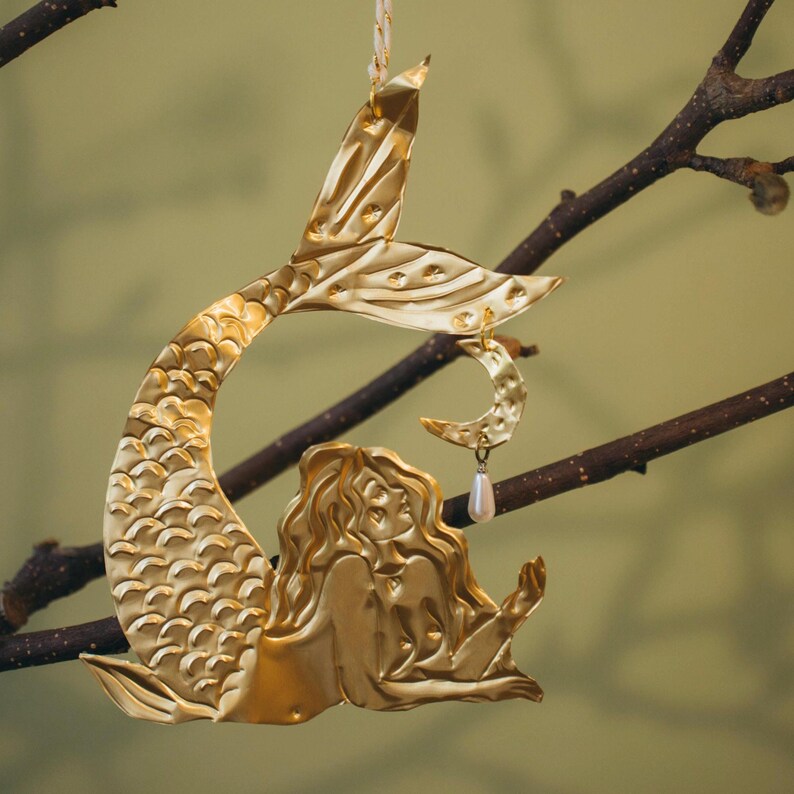 May include: A gold-colored mermaid ornament with a crescent moon and pearl charm. The mermaid is depicted with flowing hair and a detailed tail. The ornament is hanging from a branch, creating a whimsical and decorative display.