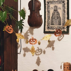 May include: A Halloween-themed garland featuring orange pumpkin cutouts, gold-colored leaves, and acorn-shaped ornaments. The garland is strung across a white wall, with a vintage violin and framed artwork in the background. The overall aesthetic is autumnal and festive.