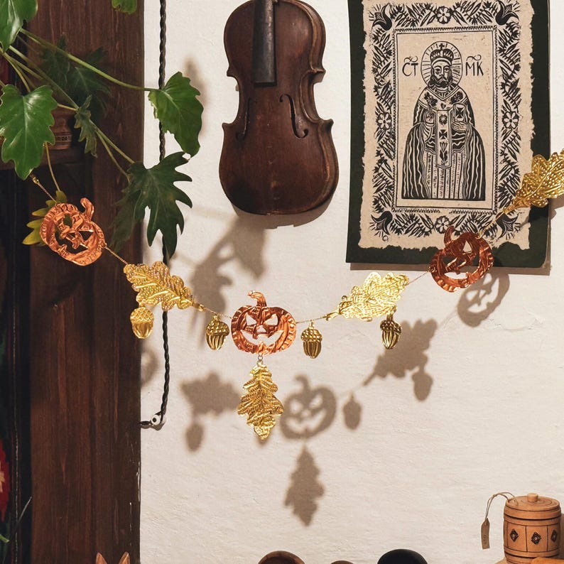 May include: A Halloween-themed garland featuring orange pumpkin cutouts, gold-colored leaves, and acorn-shaped ornaments. The garland is strung across a white wall, with a vintage violin and framed artwork in the background. The overall aesthetic is autumnal and festive.
