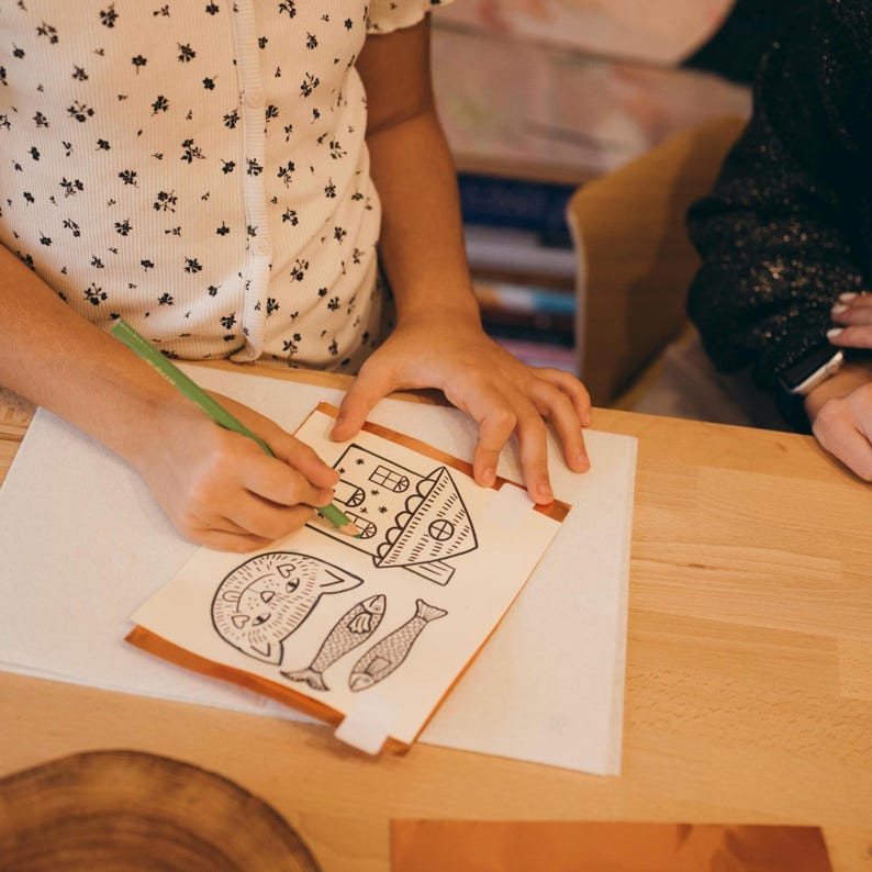 May include: A person coloring a drawing of a house, cat, and fish with a green pencil. The drawing is on a white sheet of paper with copper tape edges. The person is wearing a white shirt with a black floral pattern.
