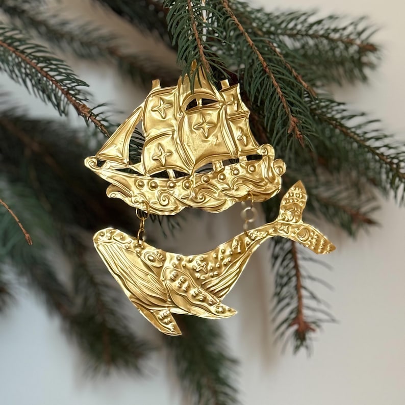 May include: Two gold-colored Christmas ornaments hanging from a pine branch. One ornament is a detailed sailboat, and the other is a whale. Both ornaments have intricate designs and are suspended by small chains.