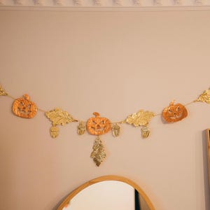 May include: A festive garland featuring orange pumpkin and gold leaf cutouts, along with acorns. The garland is strung on a thin gold cord and is hanging on a light-colored wall, perfect for autumn decor.