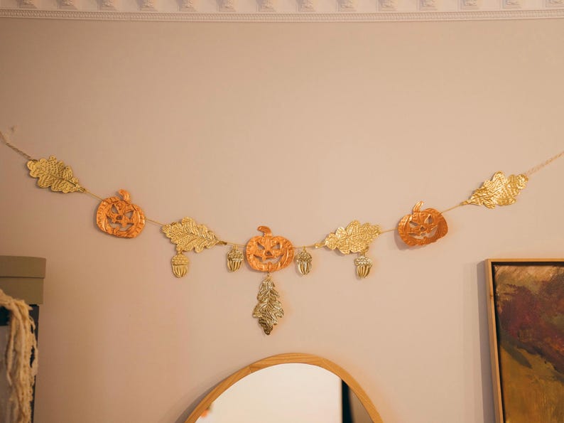 May include: A festive garland featuring orange pumpkin and gold leaf cutouts, along with acorns. The garland is strung on a thin gold cord and is hanging on a light-colored wall, perfect for autumn decor.