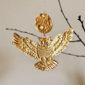 May include: A golden metal owl ornament with outstretched wings, hanging below a textured, golden moon. The owl has detailed feather patterns and is suspended by a thin wire, creating a celestial theme.