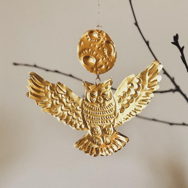 May include: A golden metal owl ornament with outstretched wings, hanging below a textured, golden moon. The owl has detailed feather patterns and is suspended by a thin wire, creating a celestial theme.