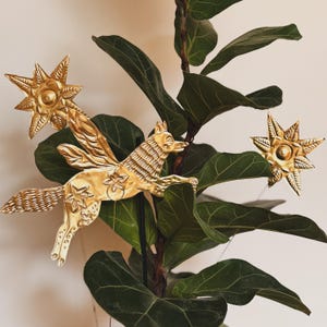 Puede incluir: Una escultura de metal dorado de un zorro alado con detalles de estrellas y dos adornos en forma de estrella. La escultura está colocada sobre un fondo de hojas verdes grandes. El zorro tiene detalles de pelaje y alas.