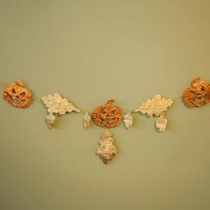 May include: A festive garland featuring orange jack-o'-lanterns, gold-colored oak leaves, and acorns. The autumn-themed decoration is strung on a thin, natural-colored cord, perfect for fall and Halloween decor.