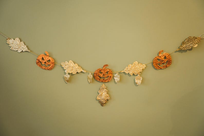 May include: A festive garland featuring orange jack-o'-lanterns, gold-colored oak leaves, and acorns. The autumn-themed decoration is strung on a thin, natural-colored cord, perfect for fall and Halloween decor.