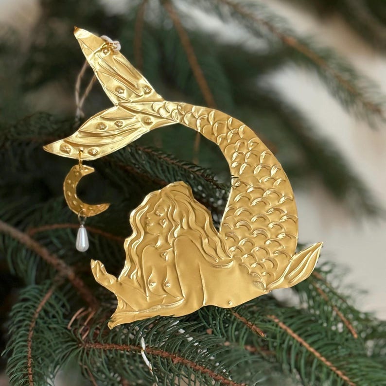 May include: A gold-colored mermaid ornament with a crescent moon and pearl charm hanging from the tail. The mermaid is lying down with flowing hair and a detailed tail. The ornament is hanging on a green Christmas tree.
