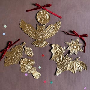 May include: Gold-colored metal ornaments in various shapes, including an owl, bat, leaf, mushroom, acorn, moon, and star. Each ornament has a textured surface and is adorned with a red ribbon for hanging. The ornaments are arranged on a brown background.