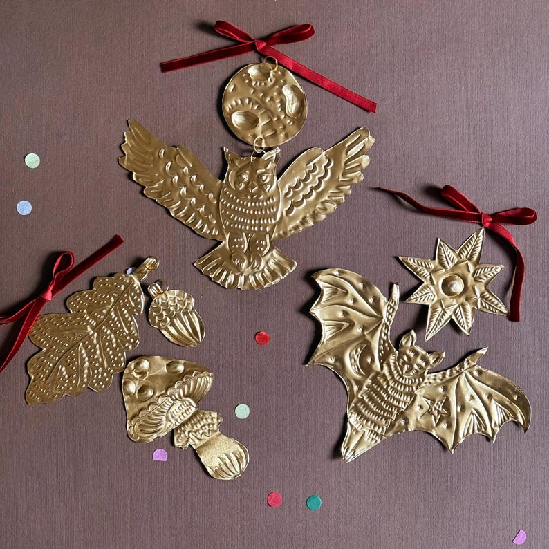 May include: Gold-colored metal ornaments in various shapes, including an owl, bat, leaf, mushroom, acorn, moon, and star. Each ornament has a textured surface and is adorned with a red ribbon for hanging. The ornaments are arranged on a brown background.