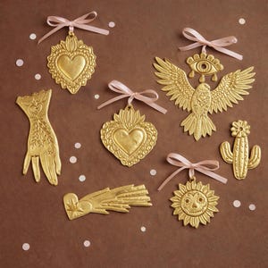 May include: A collection of gold-colored ornaments with pink ribbons. The ornaments include a heart, an eye, a hand, a cactus, a sun with a skull face, a comet, and a bird with outstretched wings. The ornaments are on a brown background with white dots.