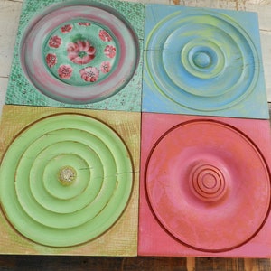 May include: Four square wooden wall decor pieces with different circular patterns. The top left piece is green with a pink floral pattern. The top right piece is blue with a concentric circle pattern. The bottom left piece is green with a concentric circle pattern. The bottom right piece is red with a concentric circle pattern.