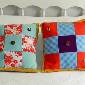May include: Two square patchwork cushions with yellow trim. One cushion is red, blue, and purple with a white and blue checked pattern. The other cushion is red, white, and blue with a floral pattern. Both cushions have buttons.