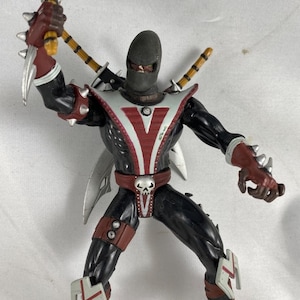 May include: Action figure of a masked character in a black suit with red and white accents. The figure has a skull belt buckle, spiked gauntlets, and two swords on its back. It is holding a weapon with a silver blade.