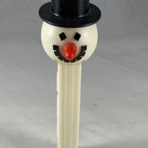 May include: A vintage PEZ dispenser shaped like a snowman. The snowman has a black top hat, a white head, and a red nose. The dispenser's body is off-white with vertical ridges.