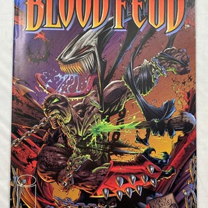 May include: A comic book titled Spawn Blood Feud issue 2 featuring a colorful illustration of a dark, muscular character with chains, a spiked red gauntlet, and a creature with sharp teeth and a long tongue, set against a purple and orange background.