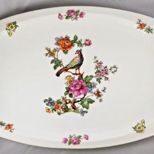 May include: An oval white ceramic serving platter featuring a central illustration of a colorful bird perched on a flowering branch with pink, orange, and yellow blossoms. Smaller floral clusters are scattered around the rim of the platter.