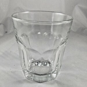 May include: A clear, faceted glass tumbler with a thick base. The glass features decorative heart-shaped indentations along the sides and a hexagonal base. The rim is smooth and the glass is empty.
