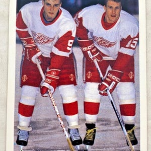May include: Two hockey players in red and white jerseys with the number 9 and 15, holding hockey sticks. The jerseys feature a winged logo. The players wear red gloves, white pants, and black skates. The card has a "90 Upper Deck 91" logo.