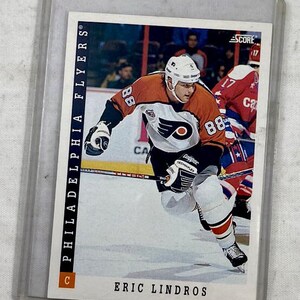 May include: A hockey trading card featuring Eric Lindros in a Philadelphia Flyers uniform. The card is in a protective sleeve. The player is wearing an orange and black jersey with the number 88. The card has the text "PHILADELPHIA FLYERS" and "SCORE".