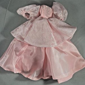 May include: A light pink doll dress with a layered design. The dress features a bodice with a hood and puffed sleeves. The skirt has a flowing, layered appearance, creating a delicate and feminine look. The fabric has a slight sheen.