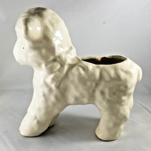 May include: A cream-colored ceramic planter shaped like a standing lamb. The surface features a distinct crackle glaze finish and a textured, curly coat pattern. The opening for plants is located on the back of the animal figure.