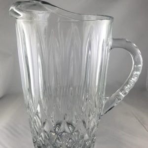 May include: A clear glass pitcher with a handle and a pouring spout. The pitcher has a textured, diamond-like pattern on the bottom and vertical lines on the sides. The handle is curved and attached to the side of the pitcher.
