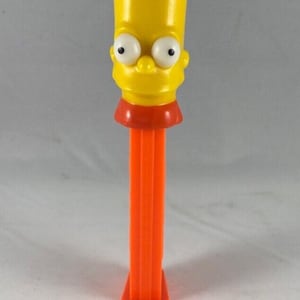 May include: A yellow Bart Simpson PEZ dispenser with a red collar and orange stem. The head features large white eyes with black pupils and a small nose. The dispenser is a collectible item.