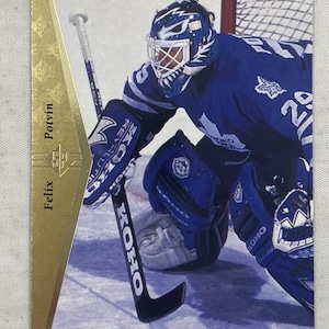Puede incluir: Una tarjeta de hockey con Felix Potvin de los Toronto Maple Leafs en uniforme de portero. La tarjeta tiene un acento de lámina dorada y el logotipo de los Maple Leafs es visible. El portero lleva un casco y protecciones azules.