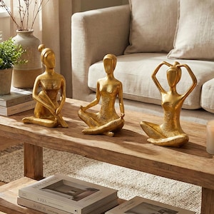 May include: Three gold-colored yoga pose figurines on a wooden coffee table. The figurines are in seated positions, with arms in various yoga poses. A lit candle and stacked books are also on the table. A vase with dried stems and a potted plant are also visible.