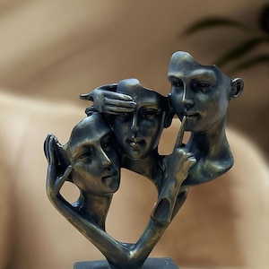 Könnte beinhalten: Bronzeskulptur, die drei menschliche Gesichter in einem stilisierten Design darstellt. Ein Gesicht bedeckt die Augen, ein anderes den Mund und das dritte das Ohr. Die Skulptur ist auf einem rechteckigen Sockel montiert.