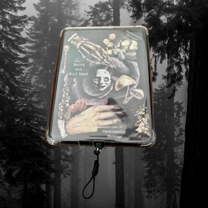 Könnte beinhalten: Eine rechteckige Handyhülle mit einem dunklen, gotischen Design. Das Kunstwerk zeigt einen Schädel, eine Skeletthand, Pilze und die Sätze "Weird and Well Read" und "Emotionally Strange". Die Hülle hat ein schwarzes Schlüsselband.