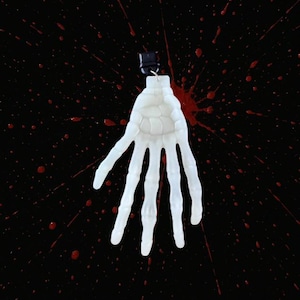 May include: A white, skeletal hand pendant with a black clasp, hanging against a black background splattered with red paint. The hand is detailed, showing the bone structure. A spooky Halloween theme.