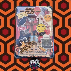 May include: A clear phone case with a collage of horror movie-themed stickers. The stickers include a crow, a smiley face, the words "LOSER", "Derry", "Carrie White Burns in Hell", and "Do Not Disturb". The case has a black border.
