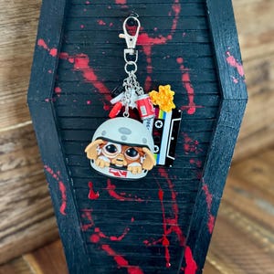 May include: A silver-toned keychain featuring a small figurine of a creature with large ears inside a white helmet, accompanied by miniature charms including a red soda can, a yellow popcorn cluster, and a black cassette tape with blue and white stripes.