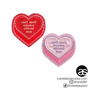 May include: Two heart-shaped embroidered patches with the text "can't spell menace without men" in white thread on a red patch and purple thread on a pink patch.