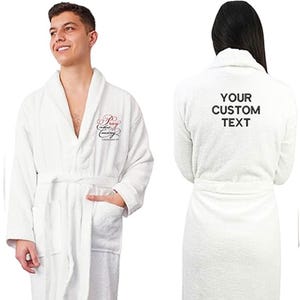 May include: White terry cloth robe with a shawl collar, pockets, and a matching belt. The front of the robe has a script design in red. The back of the robe has the text "YOUR CUSTOM TEXT" in black.