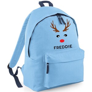 May include: A light blue backpack featuring a reindeer face design. The design includes brown antlers, black eyes, and a red nose. The name "FREDDIE" is printed in black below the reindeer face. The backpack has a front pocket and a top handle.