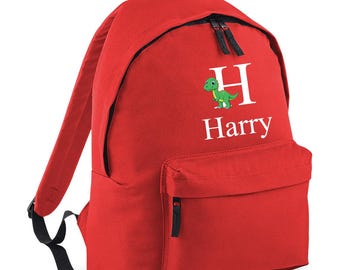 Könnte beinhalten: Ein roter Rucksack mit schwarzen Reißverschlüssen und Trägern. Der Rucksack trägt den Namen "Harry" in Weiß, einen grünen Dinosaurier und den Buchstaben "H". Er hat eine Vordertasche und einen oberen Griff.