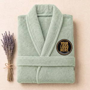 May include: A sage green terry cloth bathrobe with a shawl collar and a matching tie belt. A circular black patch is embroidered on the left chest with the gold text "YOUR LOGO HERE EMBROIDERY" in a sans-serif font. A bundle of dried lavender is nearby.
