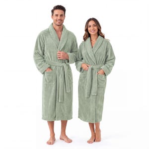Puede incluir: Dos batas de baño de felpa en color verde salvia con cuello tipo chal, mangas largas, dos bolsillos frontales y un cinturón a juego en la cintura. Las batas están confeccionadas en un tejido suave y texturizado, llegando hasta la pantorrilla.